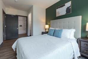 Habitación de hotel con una cama grande, cabecero, mesita de noche con lámpara y puerta que da a un baño.