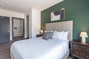 A hotel room with a large bed, a headboard, two bedside tables with lamps, a wall-mounted artwork, and a door leading to a bathroom. - Landing | Stunning 2BD, Pool, Clubhouse (Dayton)