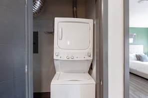 A compact, white laundry room with a built-in washer and dryer, a visible vent, and a bed with a green wall in the adjacent room. - Landing | Stunning 2BD, Pool, Clubhouse (Dayton)