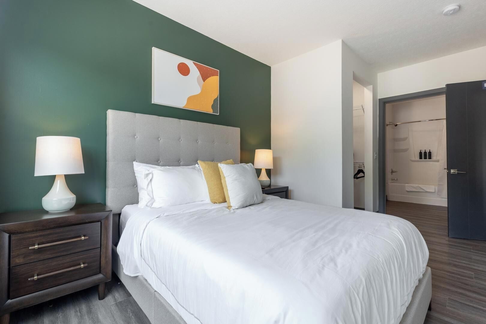 Une chambre d’hôtel avec un grand lit, deux tables de chevet avec des lampes, une tête de lit, une œuvre d’art murale et un dressing.