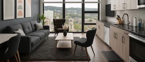 Una sala moderna con sofá, mesa de centro y sillas, una cocina con armarios y electrodomésticos, y ventanas amplias con vista a la ciudad.