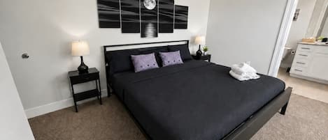Un dormitorio con una cama grande, dos mesitas de noche, una obra de arte en la pared y una puerta a otra habitación.