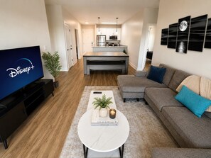 Ein modernes Wohnzimmer mit einem Flachbildfernseher, einem Couchtisch mit Büchern und einer Pflanze, einem grauen Sofa und einer Küchenzeile im Hintergrund.