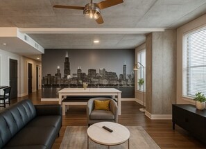 A modern living room with a cityscape mural, a ceiling fan, a sofa, a coffee table, and a dining area. - Trendy 3BR/3BA City Apt– Pool+Ping Pong 5F (Birmingham)