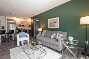 A modern living room with a grey sofa, a glass coffee table, and a white armchair. There is a kitchen visible in the background. - Landing | Incredible 2BD, Clubhouse, Courtyard (Dayton)