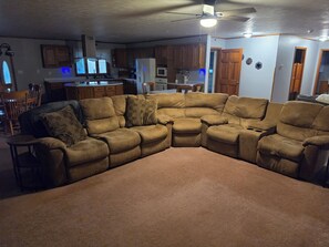 Ein Wohnzimmer mit einem Ecksofa, einem Deckenventilator und einer Küche im Hintergrund.