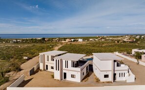 A modern residential area with white buildings, a clear view of the ocean, and a sandy landscape. - Dreamy desert villa: luxe pool & ocean views (Todos Santos)