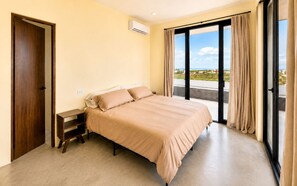 A bedroom with a bed, a nightstand, an air conditioning unit, and a view through a large window. - Dreamy desert villa: luxe pool & ocean views (Todos Santos)