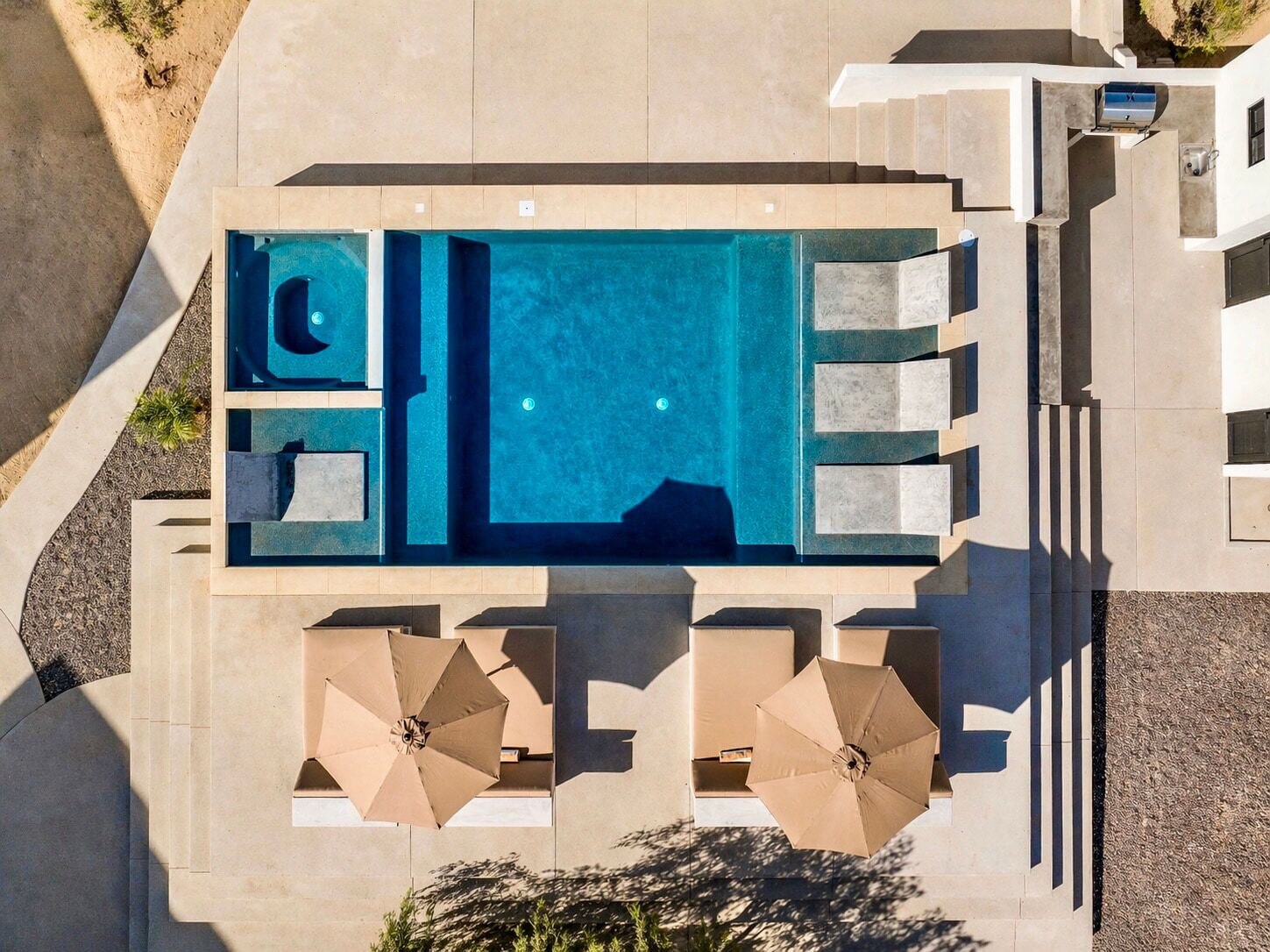 An aerial view of a rectangular swimming pool with a built-in jacuzzi, surrounded by lounge chairs and umbrellas, set against a modern architectural backdrop.