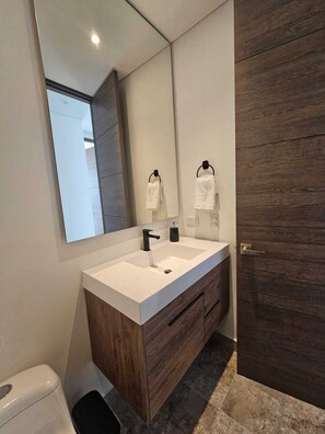 Ein modernes Badezimmer mit einem weißen Waschtisch, dunklen Holzschränken, einem großen Spiegel und einer Holztür.