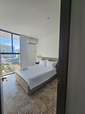 Ein Schlafzimmer mit einem großen Bett, einem Nachttisch, einer Wand-Klimaanlage und Blick auf die Stadt durch ein großes Fenster.