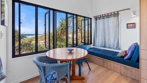Una habitación con un ventanal grande que ofrece vistas a la playa, una mesa redonda de madera, sillas de mimbre, un banco con cojines y un ventilador de techo.