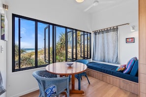 A room with a large window offering a view of the beach, a round wooden table, wicker chairs, a bench with cushions, and a ceiling fan.