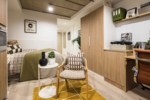 A compact living space with a bed, a small table with a plant, a chair, and a wardrobe. - Arthur House (Wembley)