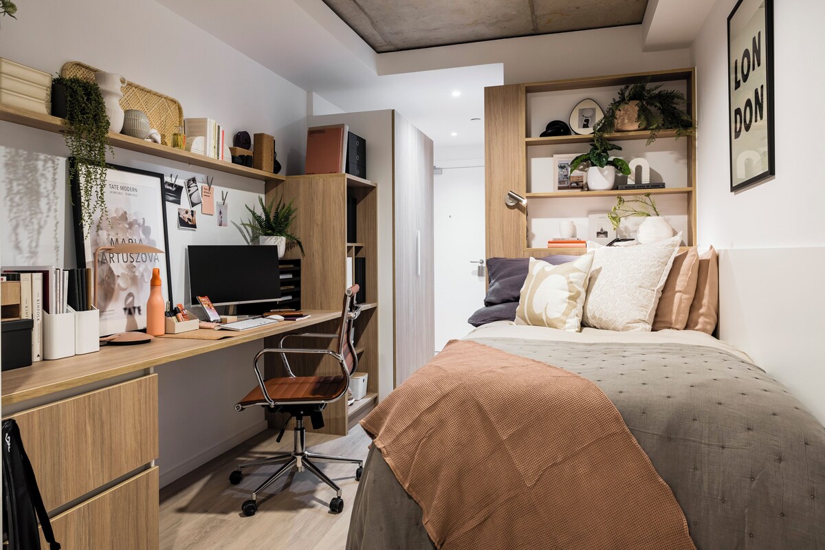A modern bedroom with a bed, desk, chair, and shelving with books and decorative items.