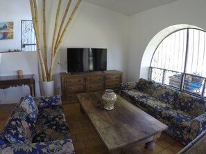 A living room with a wooden coffee table, floral sofas, a television on a cabinet, and a large arched window.