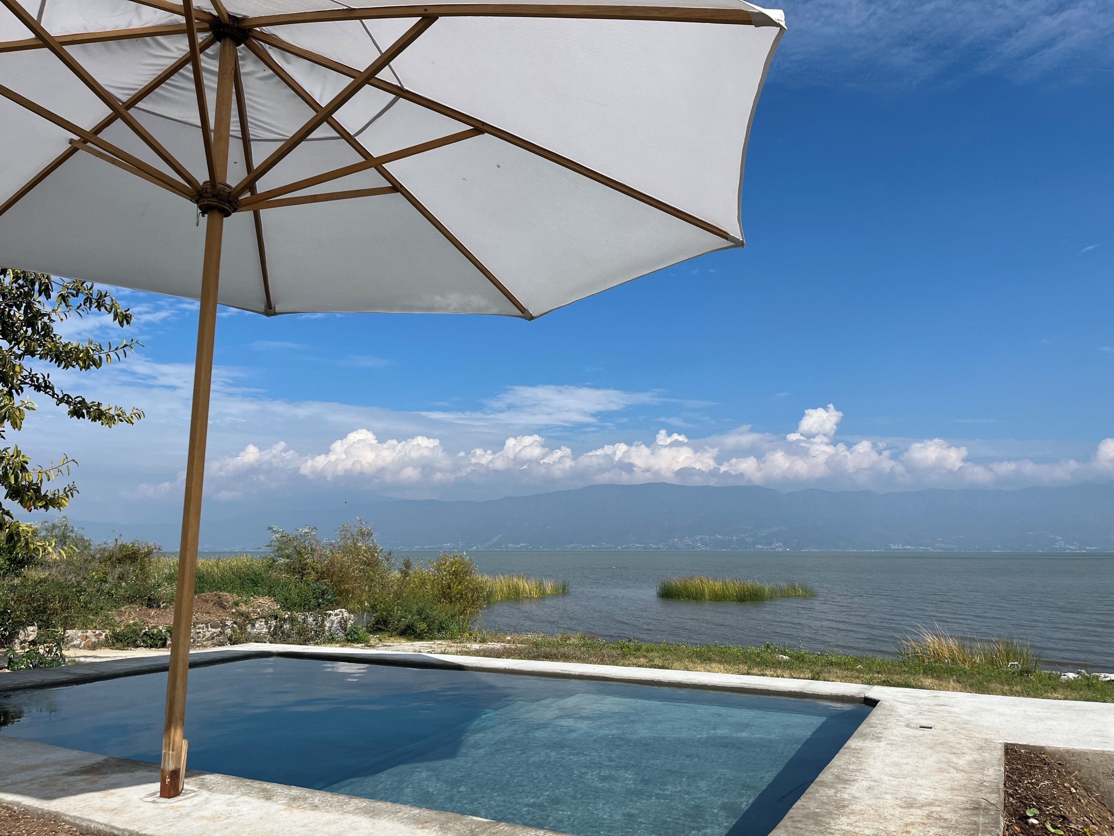 Piscina translúcida com guarda-sol de madeira, cercada por vegetação e com vista para um corpo d'água com montanhas ao longe.