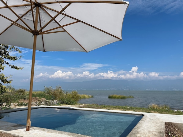 Une piscine aux eaux cristallines, agrémentée d’un parasol en bois, entourée de verdure et offrant une vue sur un plan d’eau, avec des montagnes en toile de fond.