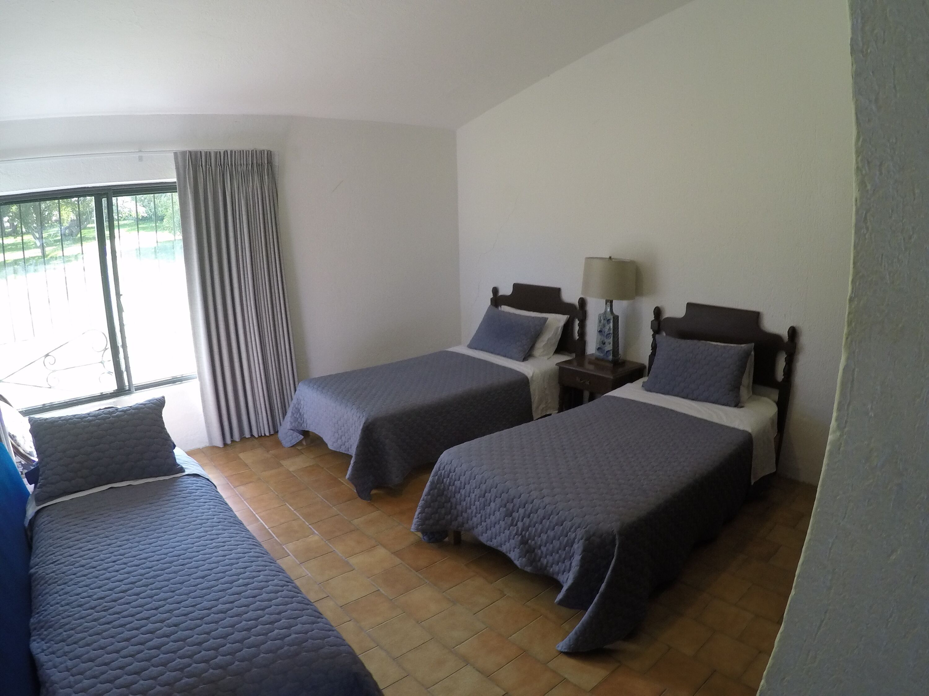 Quarto de hotel com duas camas de solteiro, mesa de cabeceira com abajur, janela com cortinas e vista para o exterior.