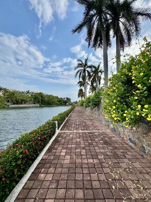 A paved walkway beside a body of water, lined with palm trees and flowering plants. - Villa Coco - Private Pool, Ocean View, and Ixtapa Marina (Zihuatanejo de Azueta)