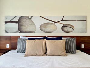 A bed with a textured white cover, two beige pillows, and two striped pillows. Above the bed, there is a wall-mounted artwork featuring three stones and branches. - Villa Coco - Private Pool, Ocean View, and Ixtapa Marina (Zihuatanejo de Azueta)