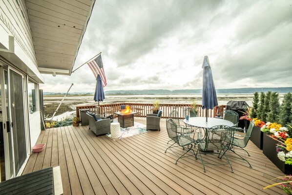 A wooden deck with outdoor furniture, a fire pit, and a view of the beach.