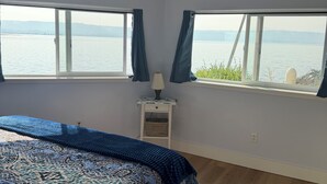 Un dormitorio con una cama grande, dos ventanas con cortinas, una mesita de noche con una lámpara y vista a un cuerpo de agua.