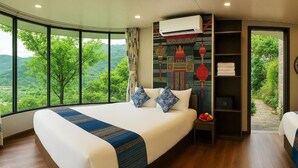 Une chambre d’hôtel avec un grand lit, une table de chevet, une étagère avec des serviettes et une vue sur la verdure grâce à de grandes fenêtres.