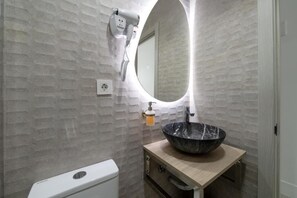 A bathroom with a marble sink, a round mirror, a hairdryer mounted on the wall, and a soap dispenser. - Rosales Suites (Madrid)