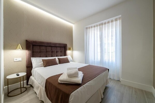 A hotel room with a large bed, a bedside table, a lamp, and a window with curtains. - Rosales Suites (Madrid)