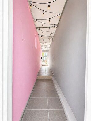 Ein enger Korridor mit einer rosafarbenen Wand auf der einen Seite und einer grauen Wand auf der anderen, der zu einem hellen Bereich mit Tageslicht führt.