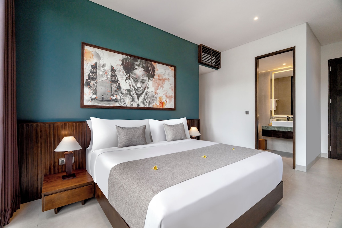 Una habitación de hotel moderna con una cama grande, dos mesitas de noche con lámparas, una obra de arte grande en la pared y un baño visible a través de una puerta abierta.