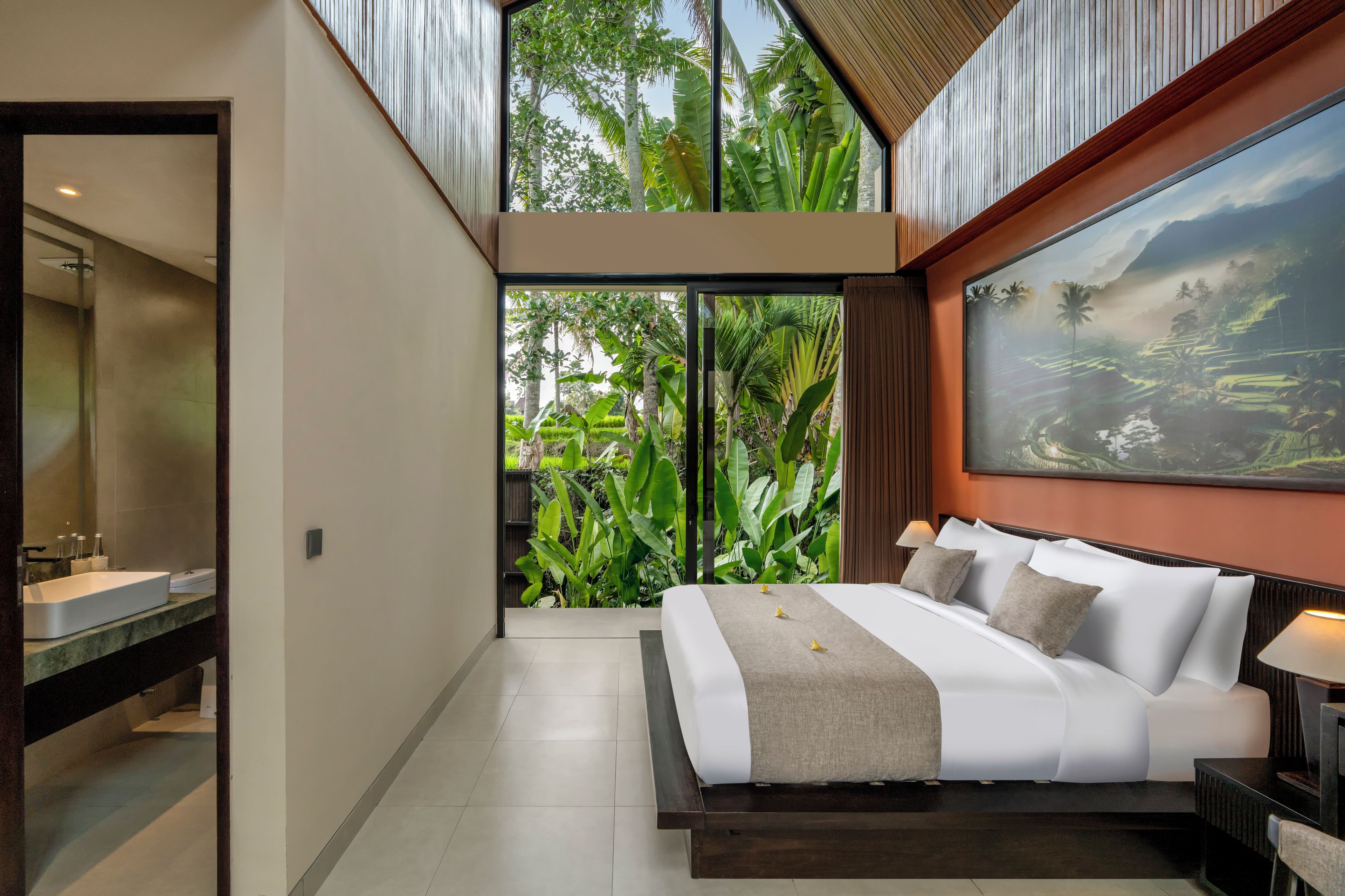 A modern bedroom with a large bed, a bathroom with a sink, and a view of lush greenery through a window.