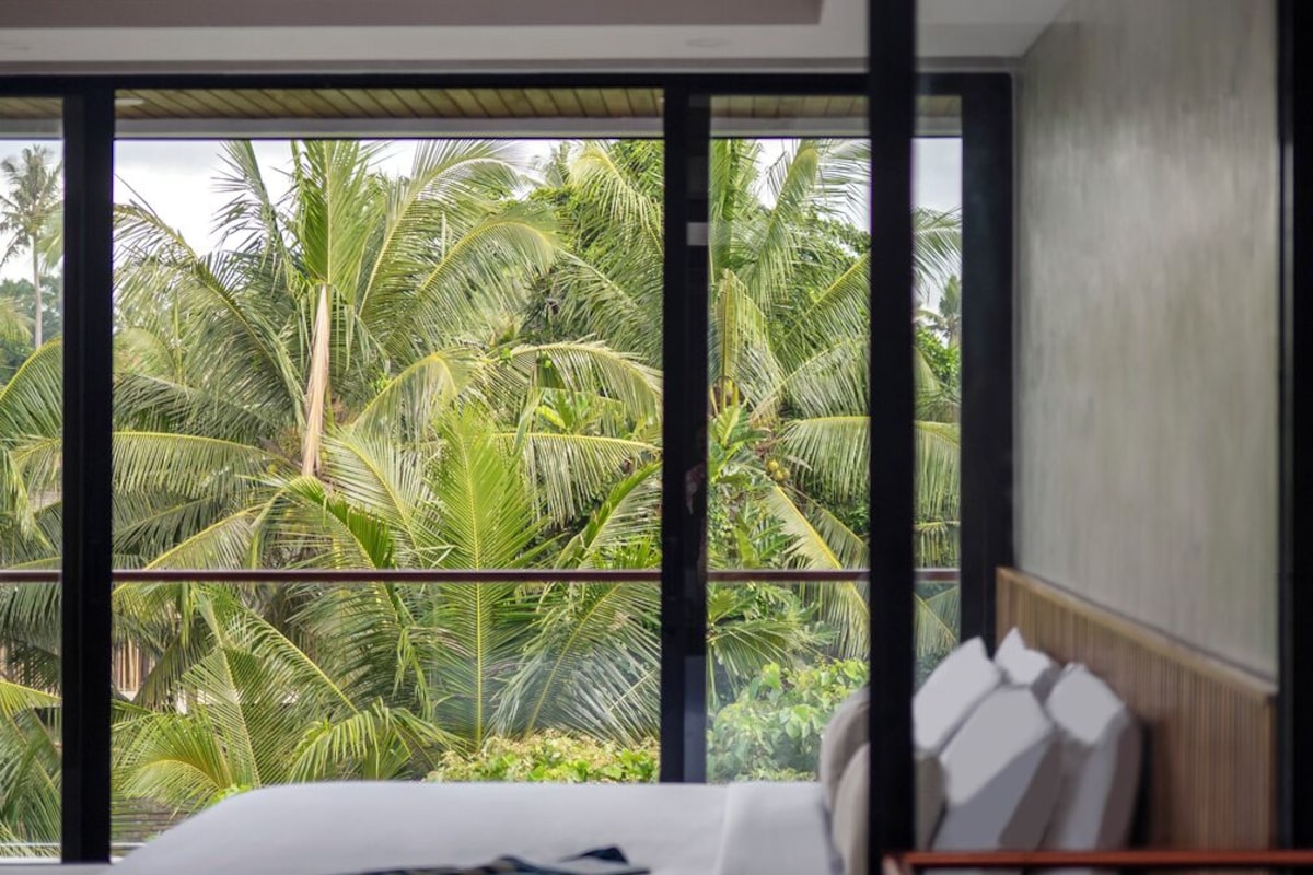 Un dormitorio con un ventanal grande, una cama con ropa de cama blanca y vistas a palmeras.