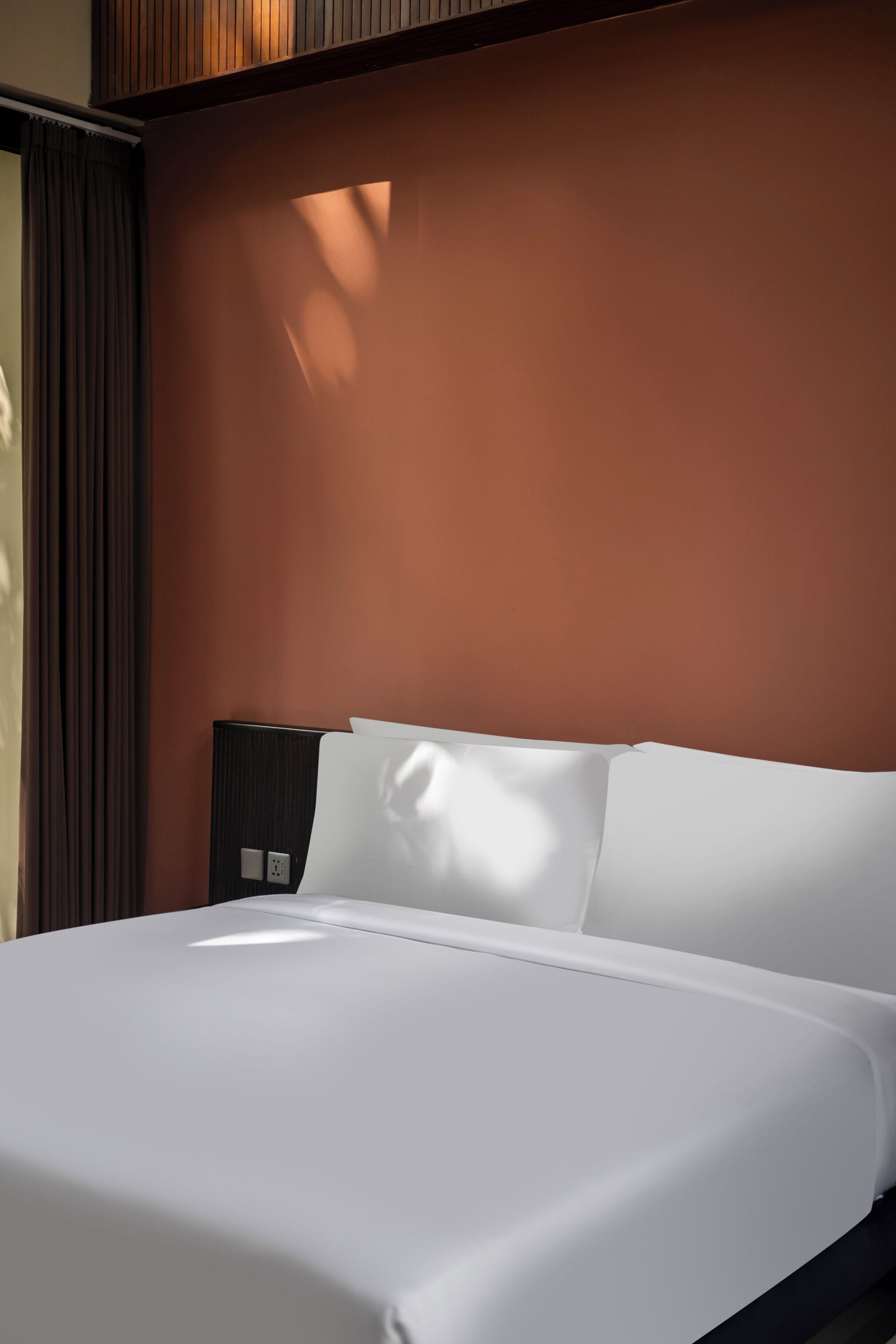 A neatly made bed with white linens in a room with a brown wall and a window casting light on the bed.