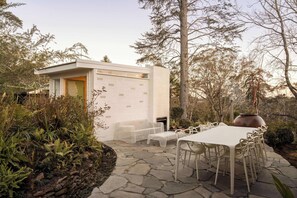 A modern outdoor patio with a stone floor, white furniture, and a small building with a door. - Sol Haus | Hot Tub, Volleyball & Fireside Nights (Wentworth Falls)