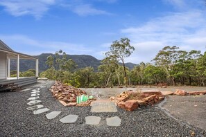 Une maison avec un chemin de pierre menant à une terrasse en bois, entourée d’un paysage naturel avec des arbres et une montagne en arrière-plan.