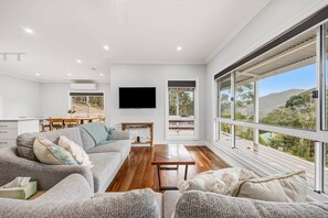 A modern living room with a grey sofa, wooden coffee table, and large windows offering a view of the outdoors. - House on the Rock | Brand New with stunning views (Halls Gap)