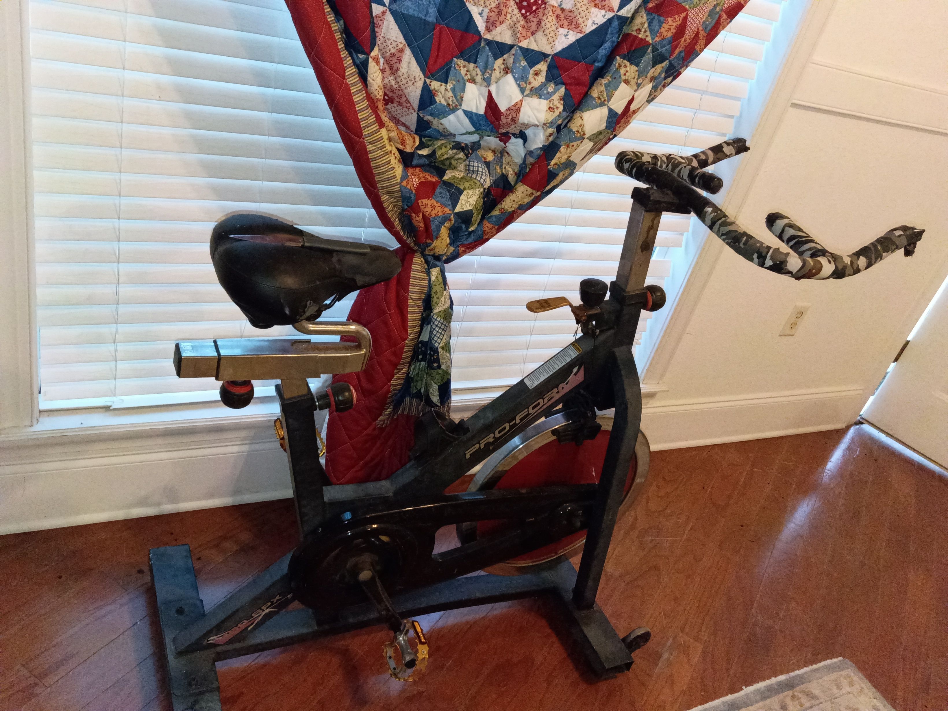 A stationary exercise bike with a red seat and handlebars, positioned in a room with wooden flooring and a window with blinds.
