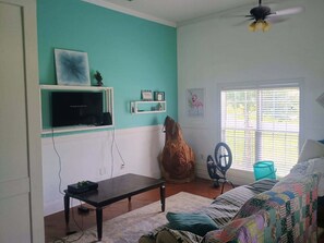 Een woonkamer met een blauwgroene wand, een houten salontafel, een bank met een kleurrijke quilt, een plafondventilator en een raam met zonwering.
