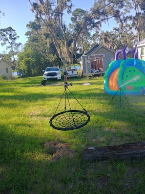 A grassy yard with a hanging net, a slide, and a small house in the background.