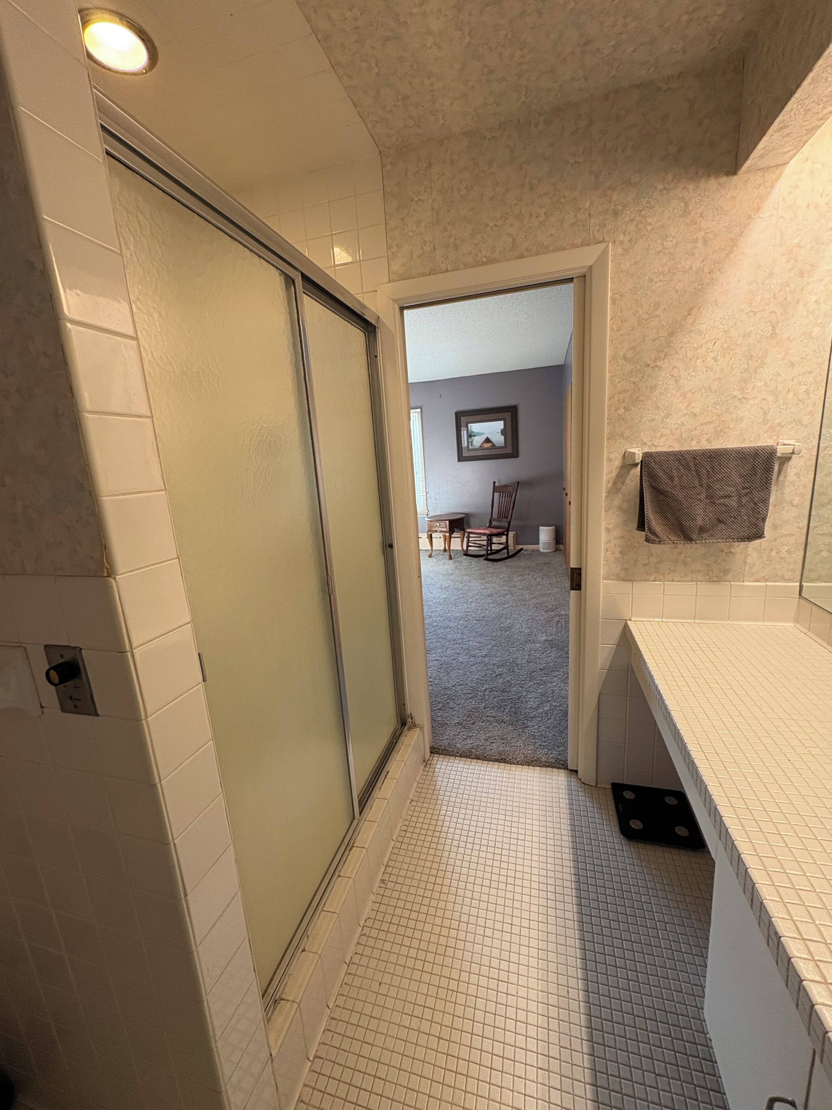 A bathroom with a tiled floor, a shower area, a sink with a towel, and a chair in the background.