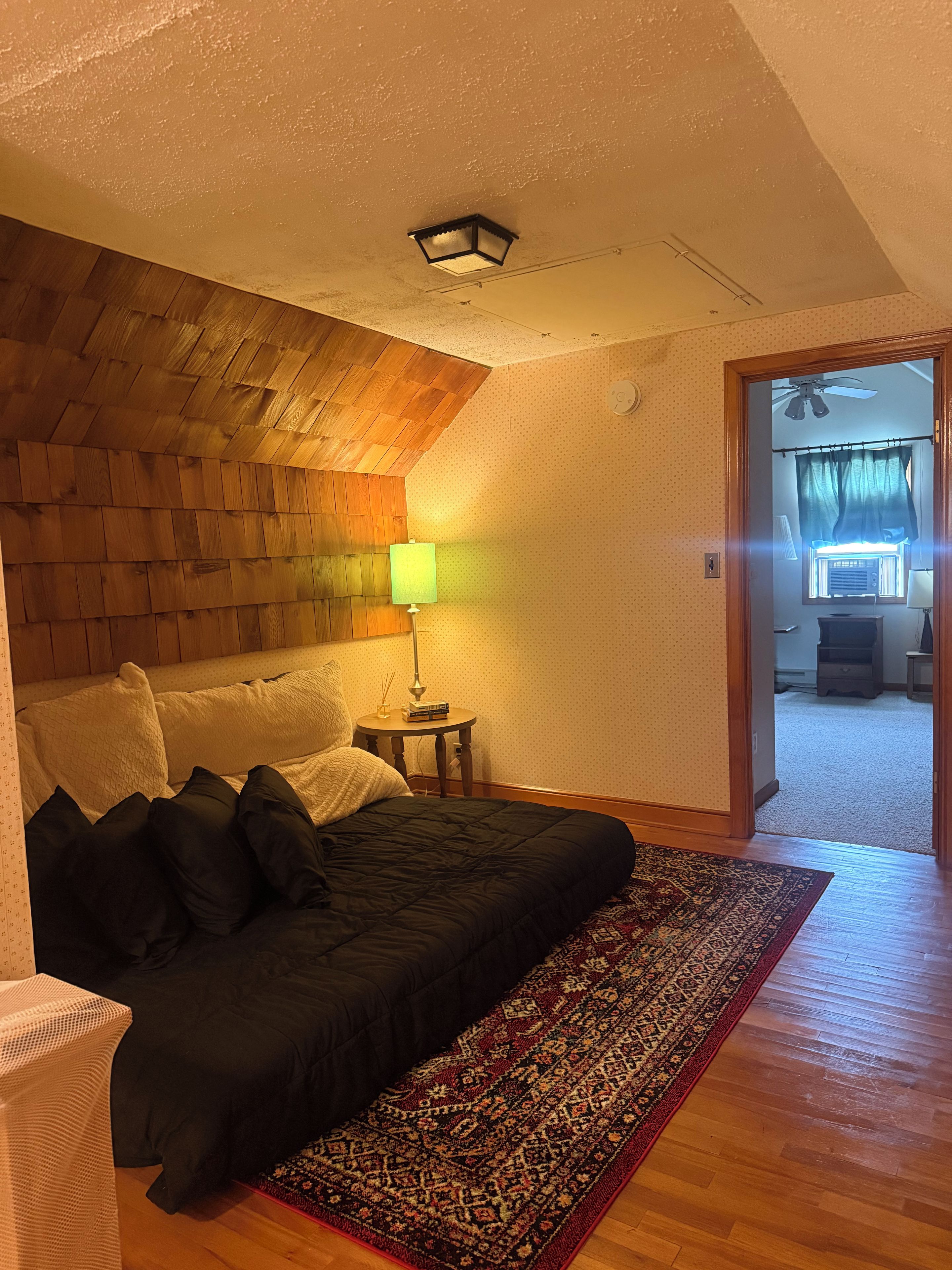 Quarto com cama, mesa de cabeceira, abajur, tapete estampado e teto de madeira.