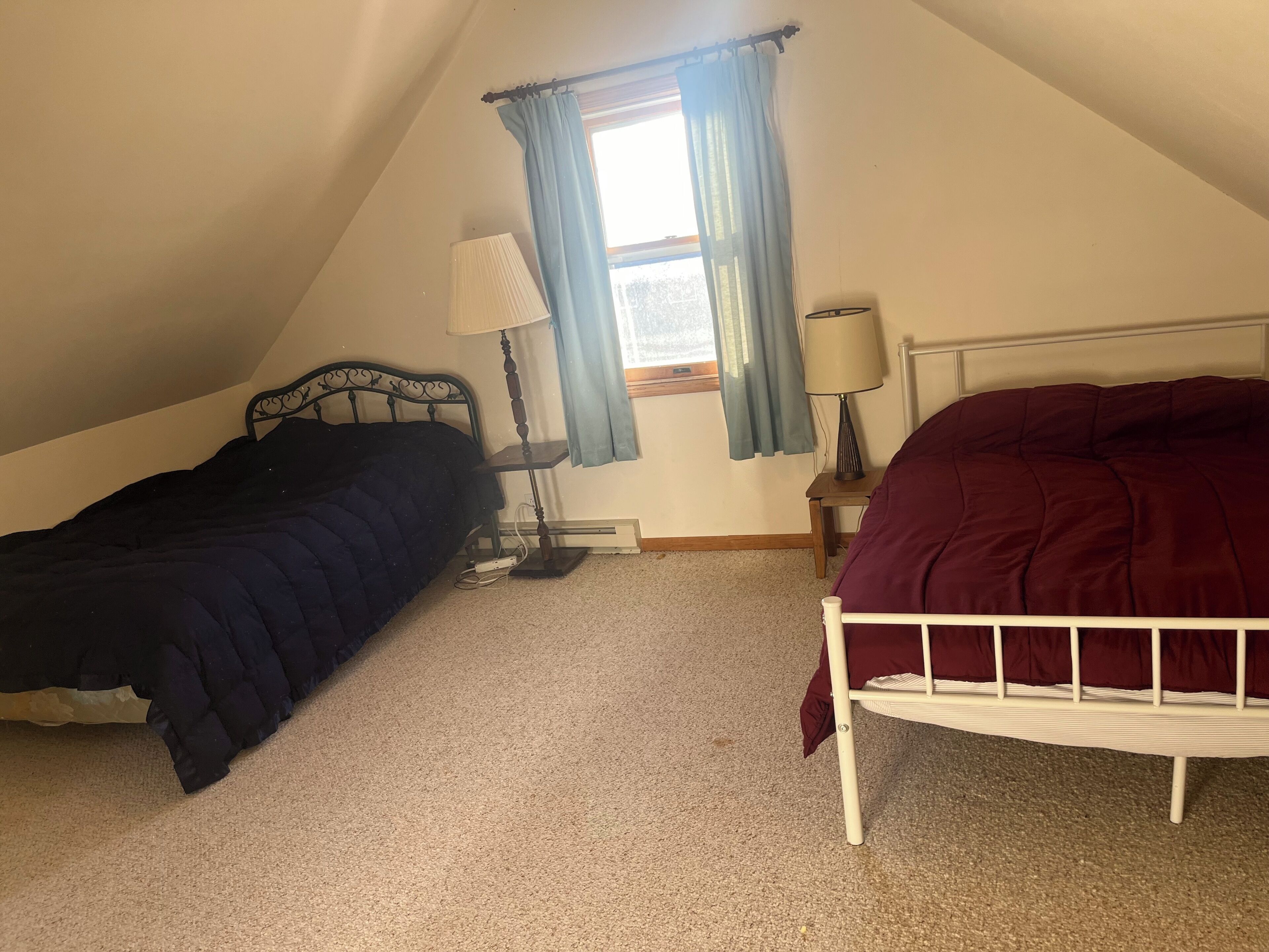 A bedroom with two beds, a window with curtains, a bedside table with a lamp, and a radiator.