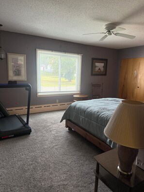 A bedroom with a bed, a treadmill, a window with blinds, a ceiling fan, and a lamp on a table.