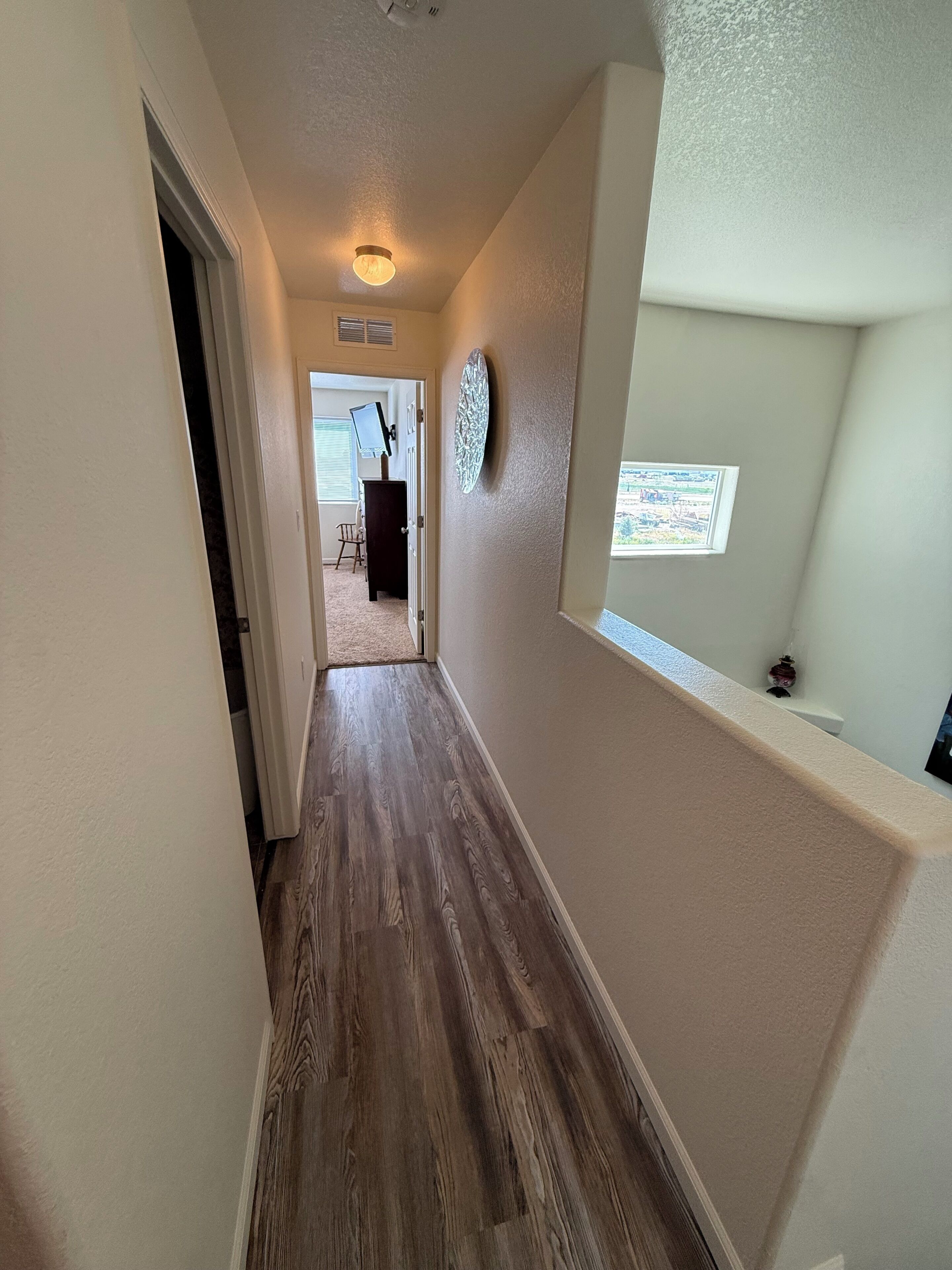 A hallway with a wooden floor, a door on the left, and a wall-mounted light fixture.