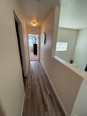 A hallway with a wooden floor, a door on the left, and a wall-mounted light fixture.