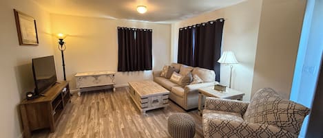 A living room with a sofa, ottoman, coffee table, TV, lamp, and curtains.