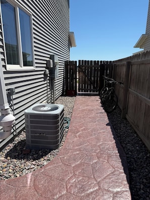 A residential outdoor area with a paved walkway, an air conditioning unit, a wooden fence, and a bicycle.
