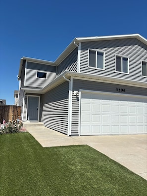 A two-story residential building with a garage, a front door, and a small front yard with grass and a wooden fence.